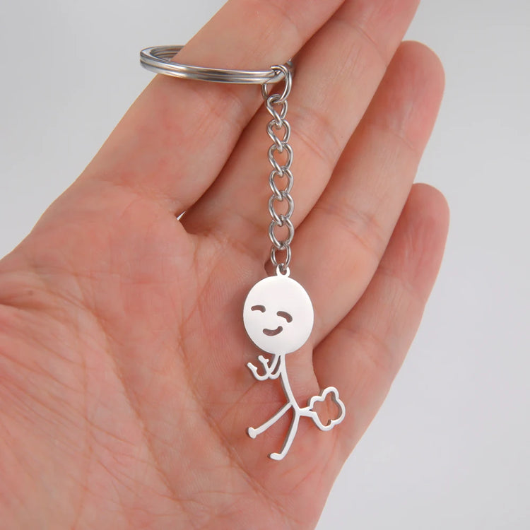 Silver keychain with a stick figure holding up 2 middle finger and farting design held in a hand against a white background
